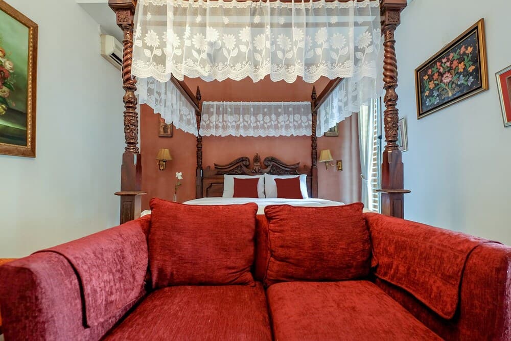 A cozy bedroom with a red velvet sofa in the foreground, a four-poster bed adorned with lace curtains, floral artwork on the walls, and warm lighting creates an inviting atmosphere ideal for a romantic getaway.