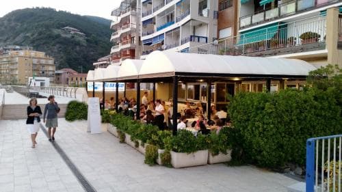 A lively seaside promenade features an outdoor café bustling with diners under white canopies, surrounded by lush greenery and modern buildings, ideal for travelers seeking a relaxing coastal dining experience.