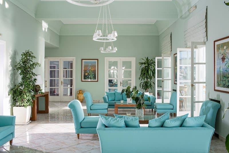 A serene hotel lobby in Thira, Greece, features light teal furnishings, lush greenery, and elegant decor, creating a tranquil atmosphere ideal for travelers seeking relaxation near cultural landmarks like the Orthodox Cathedral and Museum.
