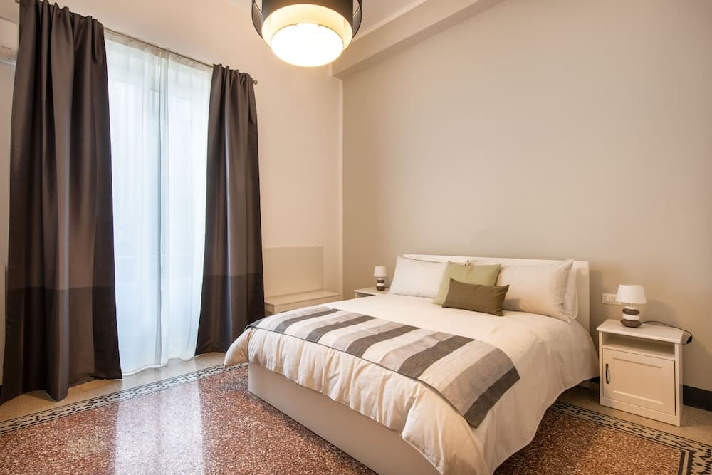 A cozy hotel bedroom features a neatly made bed with striped bedding, white nightstands with lamps, a soft glow from a ceiling light, and a window with sheer curtains, ideal for travelers seeking comfort and relaxation.
