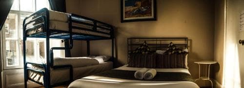 A cozy hostel room featuring bunk beds, a neatly made double bed with rolled towels, soft lighting, and framed artwork, ideal for budget travelers seeking comfort and simplicity.