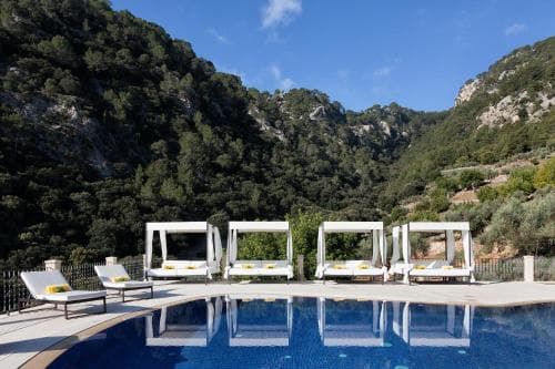 A serene luxury resort pool area surrounded by lush green mountains under a clear blue sky, featuring white lounge chairs and daybeds, ideal for a tranquil getaway.