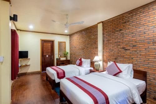 A cozy hotel room with exposed brick walls, two neatly made beds adorned with red accents, wooden furnishings, and warm lighting creates an inviting atmosphere ideal for travelers seeking comfort and charm.