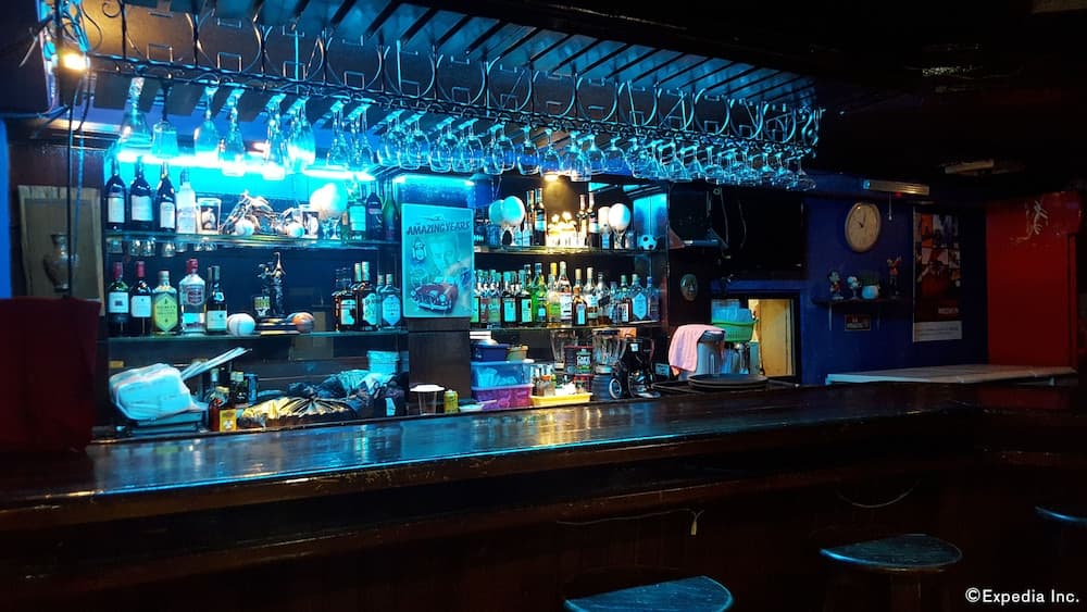 A cozy bar with a vibrant blue-lit ambiance, featuring an array of liquor bottles, hanging glassware, and eclectic decor, perfect for travelers seeking a unique nightlife experience.