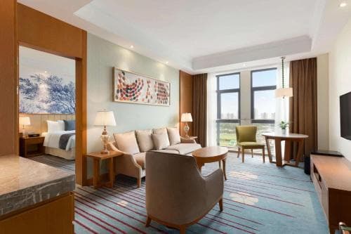 A cozy hotel suite features modern furnishings, a vibrant rug, and large windows offering a serene view, ideal for travelers seeking comfort and relaxation during their stay.