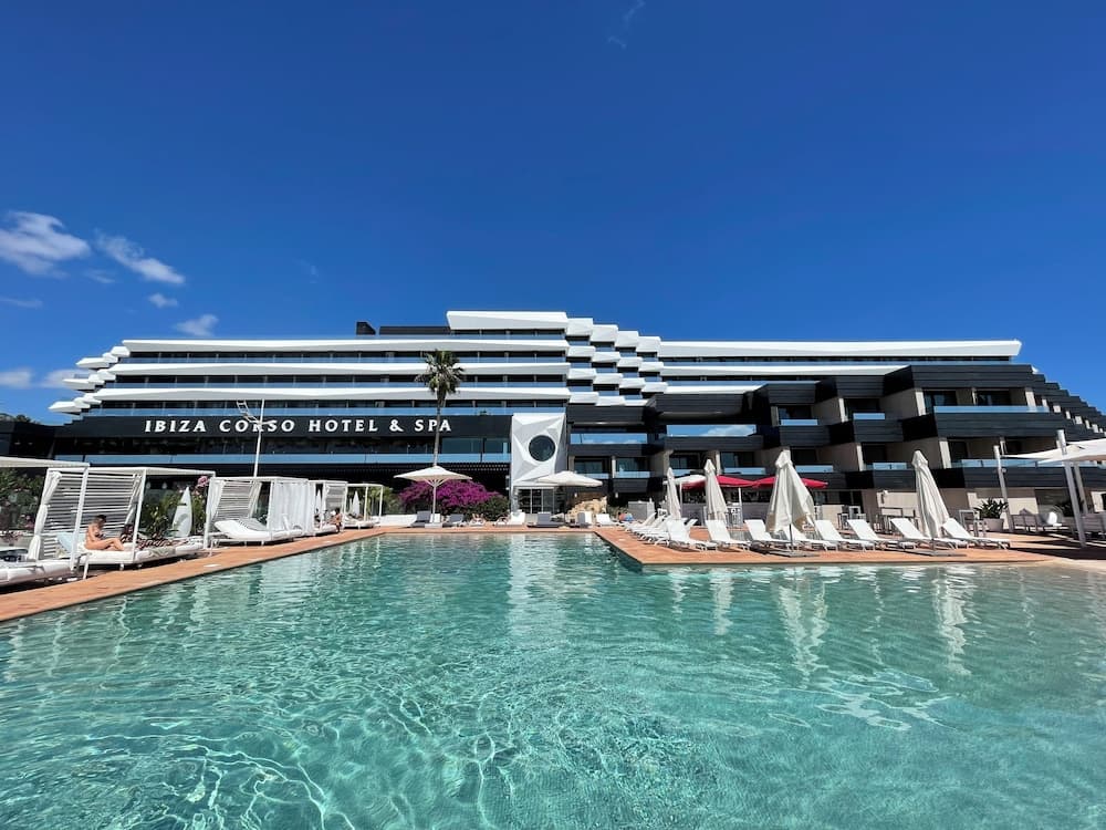 A serene luxury hotel poolside scene under a clear blue sky, featuring modern architecture, lounge chairs, umbrellas, and vibrant greenery, ideal for travelers seeking relaxation and upscale accommodations.