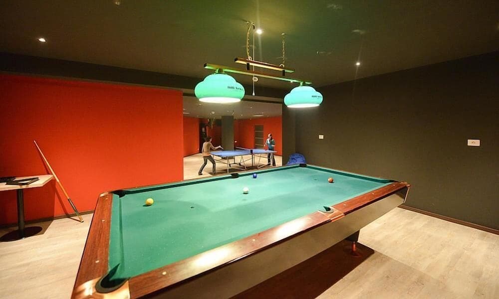 A cozy indoor recreational area features a pool table under vibrant green pendant lights, with a ping pong game in the background, ideal for travelers seeking entertainment and relaxation.