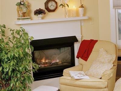 A cozy living room scene featuring a warm fireplace, plush armchair adorned with cushions and a blanket, decorative mantel with plants and ornaments, evokes a serene and inviting atmosphere perfect for relaxation or a romantic getaway.