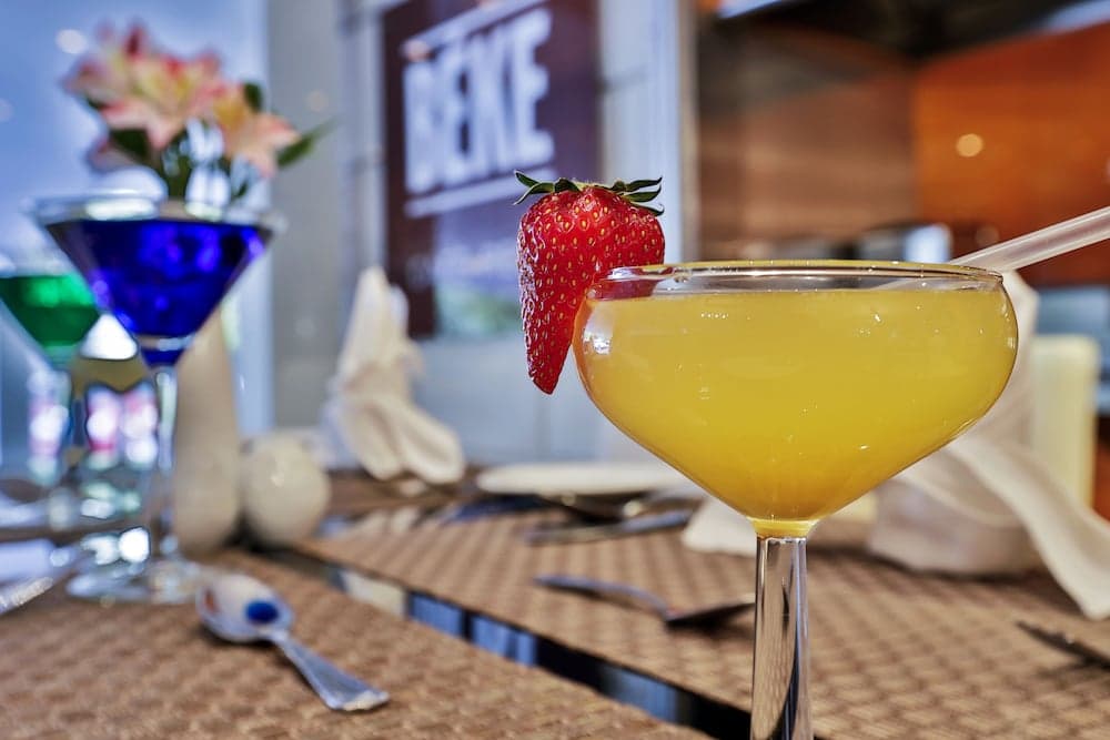 A vibrant yellow cocktail garnished with a strawberry sits invitingly on a cozy restaurant table, surrounded by colorful drinks and elegant tableware, perfect for a romantic evening out.