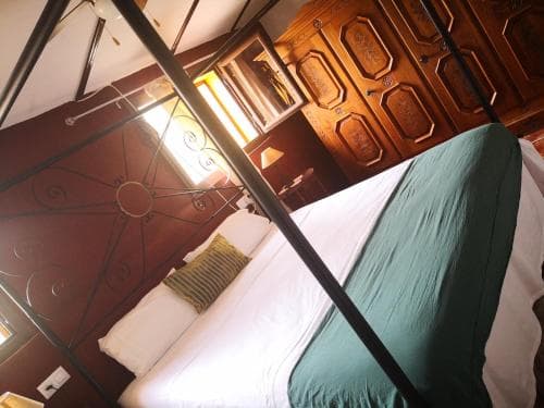 A cozy hotel room features a neatly made bed with a green blanket, ornate wooden furniture, and a decorative metal headboard, creating a warm and inviting atmosphere ideal for travelers seeking comfort.