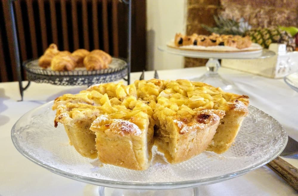 A cozy café scene features golden apple tarts on a glass stand, accompanied by croissants and a pineapple, evoking a warm, inviting atmosphere perfect for a leisurely brunch outing.