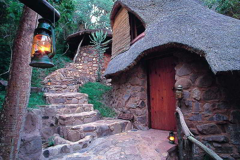 A cozy stone cottage with a thatched roof and warm lanterns invites travelers to explore its rustic charm in a serene forest setting, perfect for a tranquil getaway.