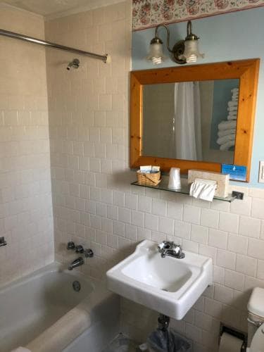 A cozy hotel bathroom features white tiled walls, a bathtub, sink, wooden-framed mirror, and neatly arranged toiletries, ideal for travelers seeking comfort and cleanliness.