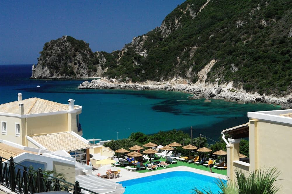 A serene coastal resort features a vibrant blue pool surrounded by lounge chairs and umbrellas, set against a backdrop of lush green cliffs and turquoise waters, ideal for a relaxing beachside getaway.