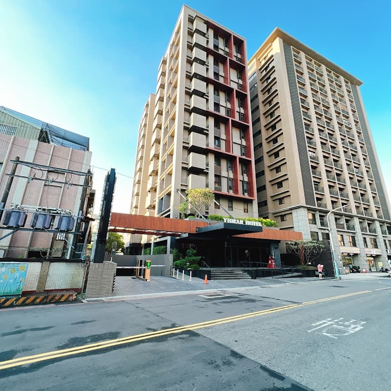A modern urban hotel entrance with sleek architecture and vibrant greenery under a clear blue sky, ideal for travelers seeking accommodation in the bustling Xinzhuang District of New Taipei City, Taiwan.