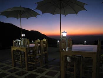 A serene outdoor dining area at dusk features wooden tables under large umbrellas, set against a breathtaking mountain sunset, ideal for romantic getaways or tranquil evening meals.