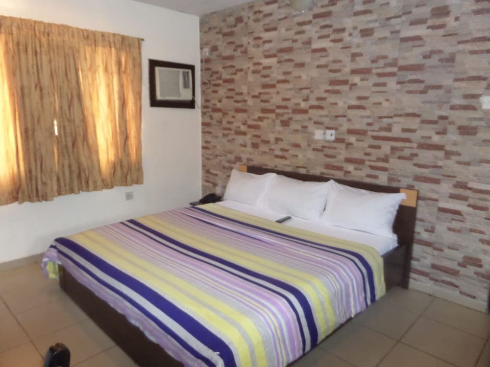 A cozy hotel room features a neatly made bed with colorful striped bedding against a textured brick wall, complemented by warm curtains and a small air conditioner, ideal for travelers seeking comfort and simplicity.