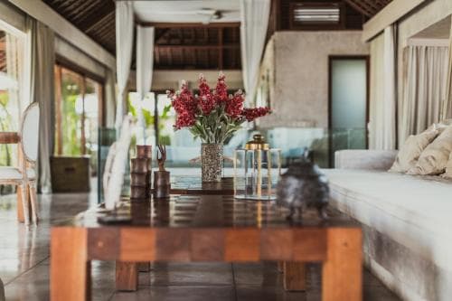 A serene tropical villa interior features a polished wooden table adorned with vibrant red flowers, inviting travelers seeking relaxation and luxury.
