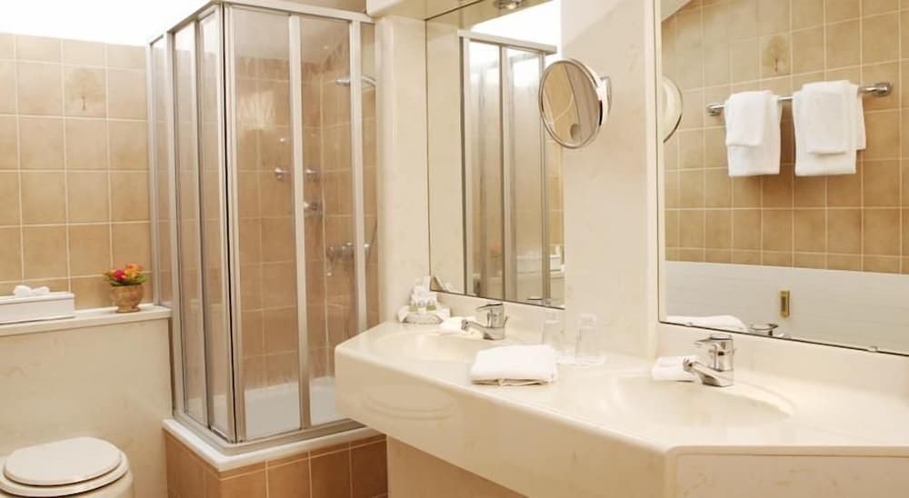 A cozy hotel bathroom features beige tiled walls, a glass-enclosed shower, a spacious sink with a large mirror, neatly folded towels, and a small vase of flowers, ideal for travelers seeking comfort and cleanliness.