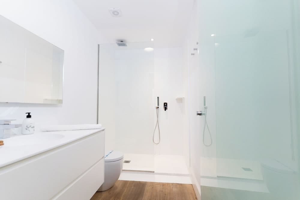 A serene modern bathroom features a minimalist white design with a glass-enclosed shower, sleek vanity, and wooden flooring, ideal for travelers seeking clean and contemporary accommodations.