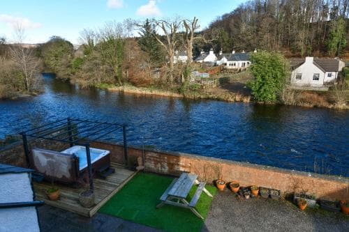 A serene riverside retreat features a cozy patio with a hot tub, picnic table, and lush greenery, ideal for tranquil vacations amidst nature.