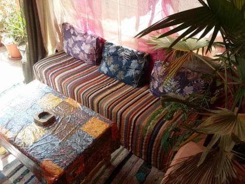 A cozy indoor lounge area features a vibrant striped sofa adorned with patterned cushions, a colorful patchwork tablecloth, and lush green plants, creating a relaxed and inviting atmosphere ideal for travelers seeking a tranquil retreat.