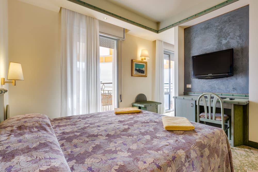 A cozy hotel room with a neatly made bed, a desk area, and a balcony offering a serene view, ideal for travelers seeking comfort and relaxation.