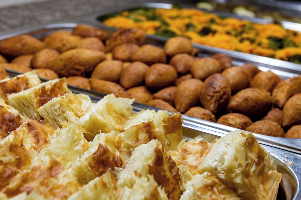 A vibrant food market display featuring golden pastries, fried snacks, and colorful salads in metal trays, inviting travelers to explore local culinary delights.