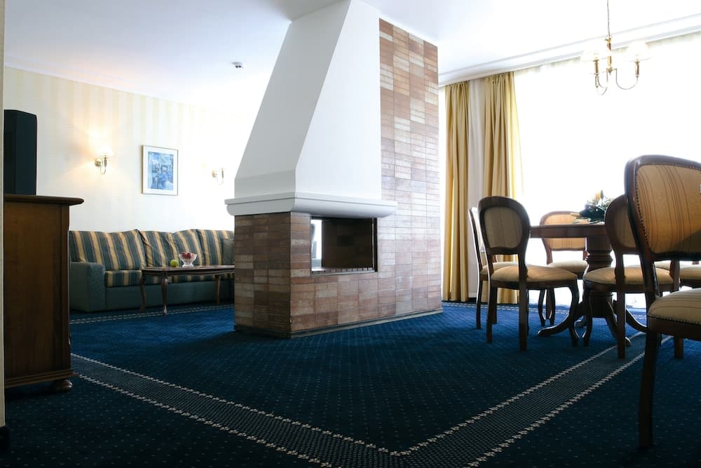 A cozy hotel lounge features a brick fireplace, striped sofa, wooden dining set by a sunlit window, and warm lighting, ideal for travelers seeking relaxation and comfort.
