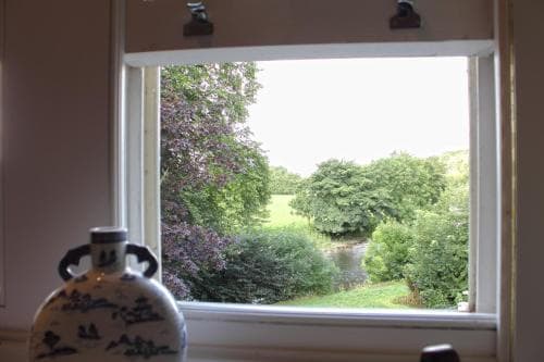 A serene countryside view framed by a window, featuring lush greenery and a tranquil stream, evokes a peaceful rural retreat, ideal for travelers seeking relaxation and nature immersion.