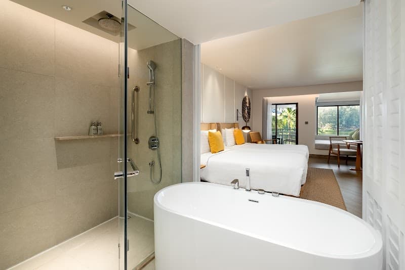 A luxurious hotel suite at SAii Laguna Phuket features a modern bathroom with a freestanding bathtub and glass-enclosed shower, leading to a cozy bedroom with a king-sized bed and tropical views, ideal for a serene and upscale vacation experience.
