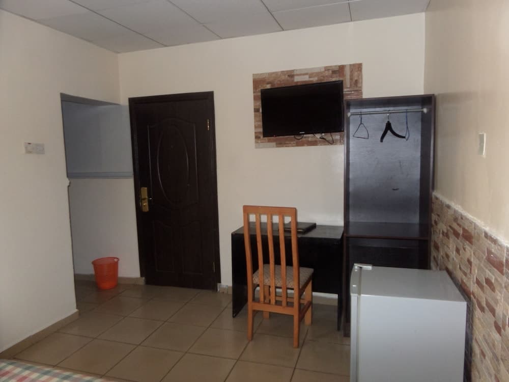 A cozy hotel room features a tiled floor, a wooden desk with chair, a mounted TV above brick-patterned walls, a small fridge, and a closet with hangers, ideal for travelers seeking comfort and functionality.