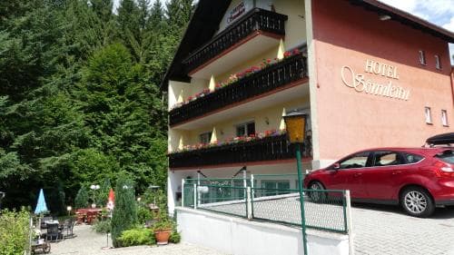 A cozy hotel nestled in a lush forest setting offers a serene retreat, featuring balconies adorned with flowers, outdoor seating, and a welcoming atmosphere ideal for travelers seeking relaxation and nature immersion.