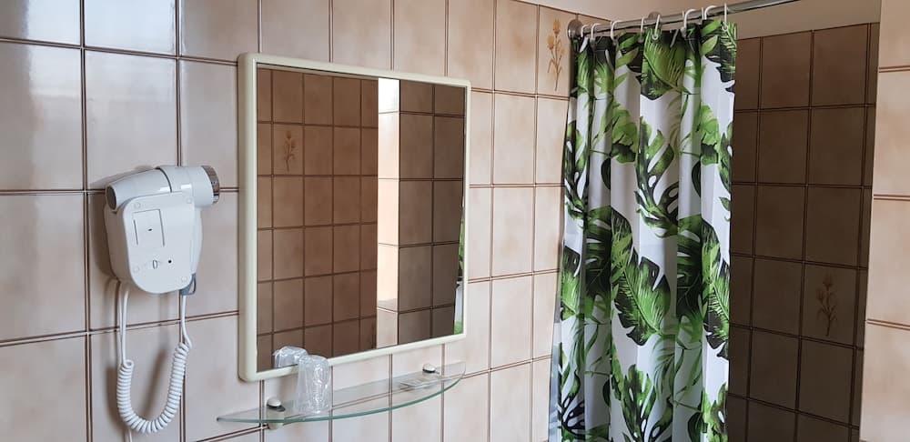 A cozy hotel bathroom features tiled walls, a wall-mounted hairdryer, a mirror with a glass shelf, and a vibrant green leaf-patterned shower curtain, ideal for travelers seeking comfort and convenience.