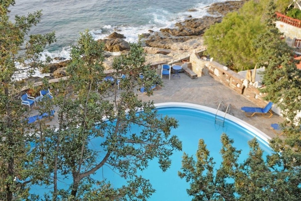 A serene coastal resort features a vibrant blue pool surrounded by lush greenery and lounge chairs, overlooking the rocky shoreline and crashing waves, ideal for travelers seeking relaxation and ocean views.