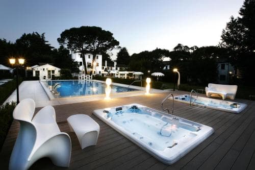 A serene evening at a luxurious resort features a glowing poolside area with modern white loungers, bubbling hot tubs, and warm lampposts, ideal for travelers seeking relaxation and upscale amenities.