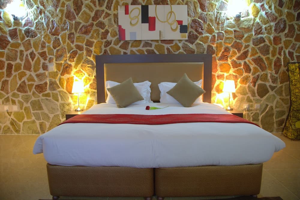 A cozy hotel room features a neatly made bed with white linens and a red accent, set against a warm, rustic stone wall illuminated by bedside lamps, ideal for travelers seeking comfort and charm.