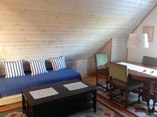A cozy attic room with wooden walls, a blue bed adorned with striped pillows, a dining table set for two, and a vibrant rug creates an inviting atmosphere perfect for a romantic getaway or a quiet retreat.