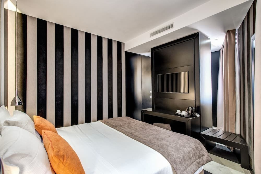 A modern hotel room features a cozy bed with orange pillows against a striking black-and-white striped wall, complemented by sleek furniture and a mounted TV, ideal for travelers seeking comfort and style.