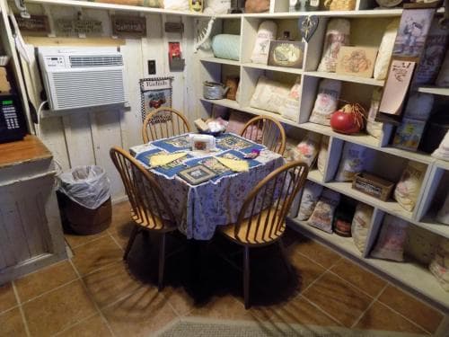 A cozy, rustic dining nook with a wooden table covered in a patchwork quilt, surrounded by shelves filled with decorative items, evokes a warm, nostalgic atmosphere perfect for a quaint getaway or a charming bed and breakfast experience.