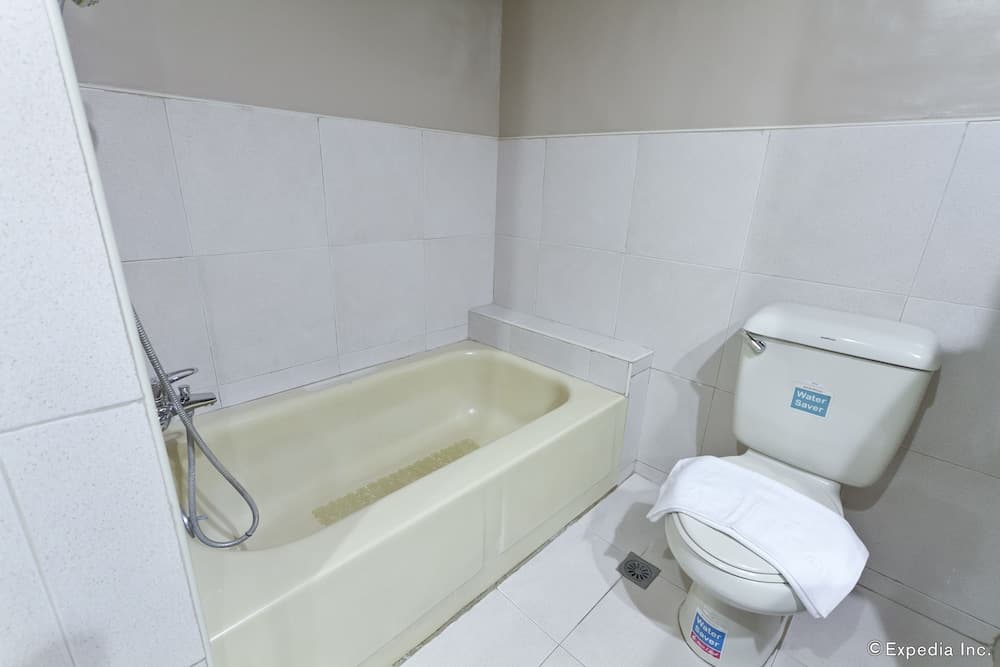 A clean, minimalist bathroom featuring a bathtub with a handheld shower, a toilet with a "Water Saver" sticker, and white tiled walls, ideal for travelers seeking basic accommodations.