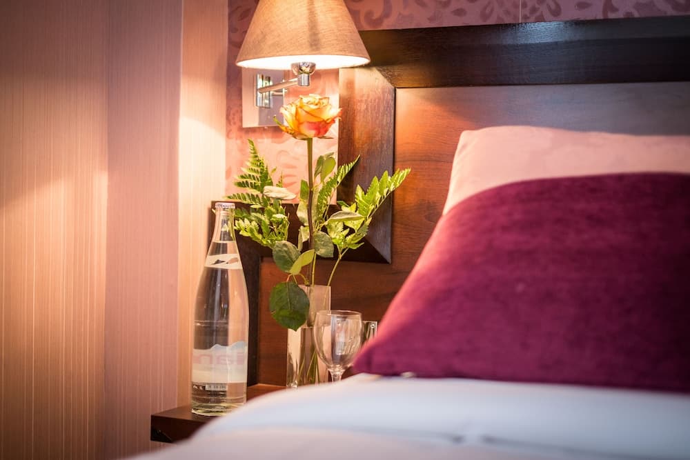 A cozy hotel room bedside scene features a warmly lit lamp, a vibrant rose in a vase, a bottle of water, and plush pillows, ideal for travelers seeking comfort and relaxation.
