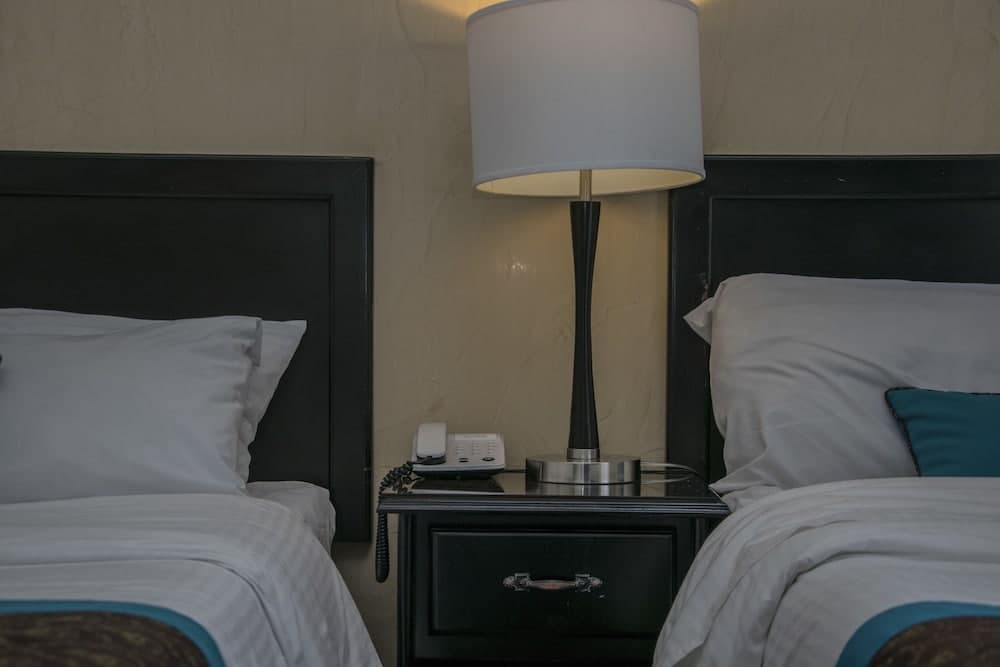 A cozy hotel room features two neatly made beds with white linens and teal accents, a central nightstand with a modern lamp and telephone, set against a textured wall, ideal for travelers seeking comfort and simplicity.