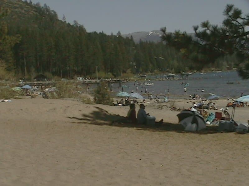 A serene lakeside beach scene with sandy shores, pine trees, and people relaxing under umbrellas, ideal for family vacations and nature enthusiasts seeking a tranquil getaway.