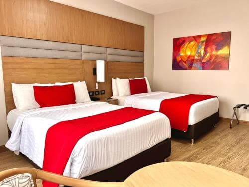 A cozy hotel room features two neatly made beds with red accents, wooden flooring, a vibrant wall art piece, and modern furnishings, ideal for travelers seeking comfort and style during their stay.