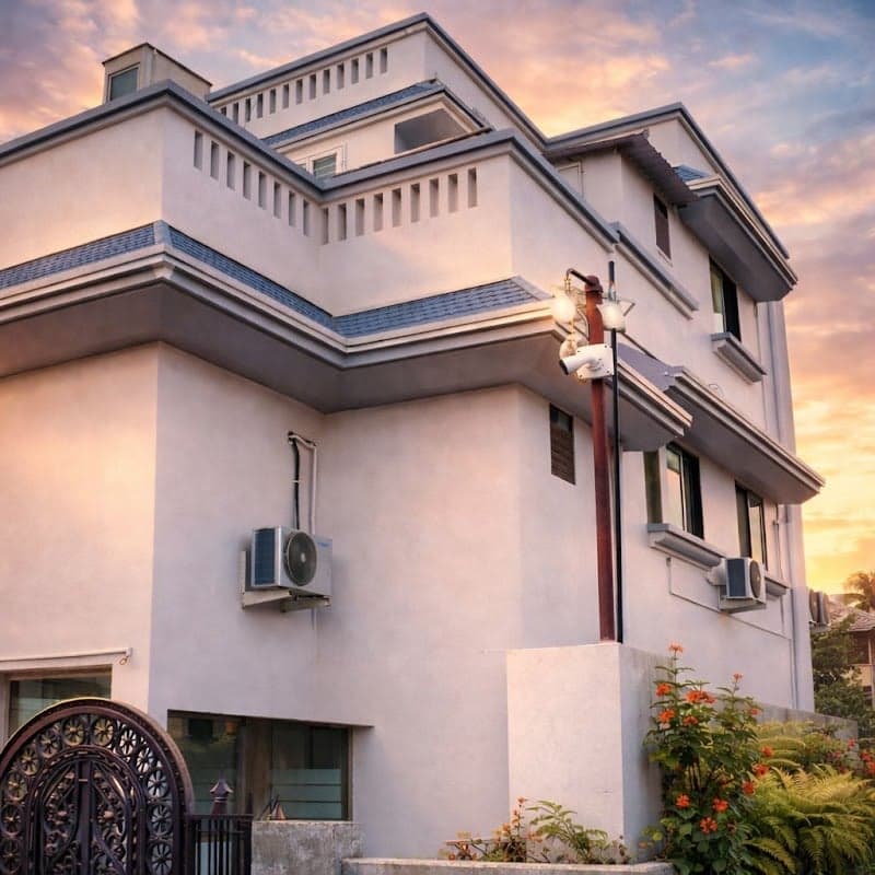 A serene evening view of a multi-story hotel in Nashik, India, featuring a decorative gate, air conditioning units, and vibrant flowers, ideal for travelers seeking accommodation in a tranquil setting.