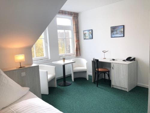 A cozy hotel room with a slanted ceiling, green carpet, white furniture, and a window view of trees, ideal for travelers seeking comfort and simplicity.