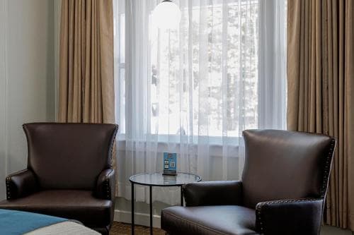 A cozy hotel room corner features two plush leather armchairs flanking a small glass table with a brochure, set against a backdrop of sheer curtains framing a snowy outdoor view, ideal for travelers seeking relaxation and comfort.