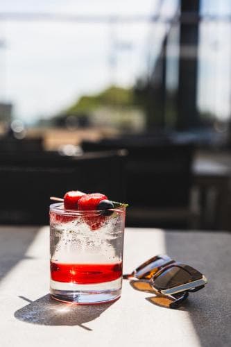 A refreshing red cocktail garnished with strawberries and mint sits on a sunlit patio table beside stylish sunglasses, evoking a relaxed summer vibe perfect for leisurely afternoons at an outdoor café.