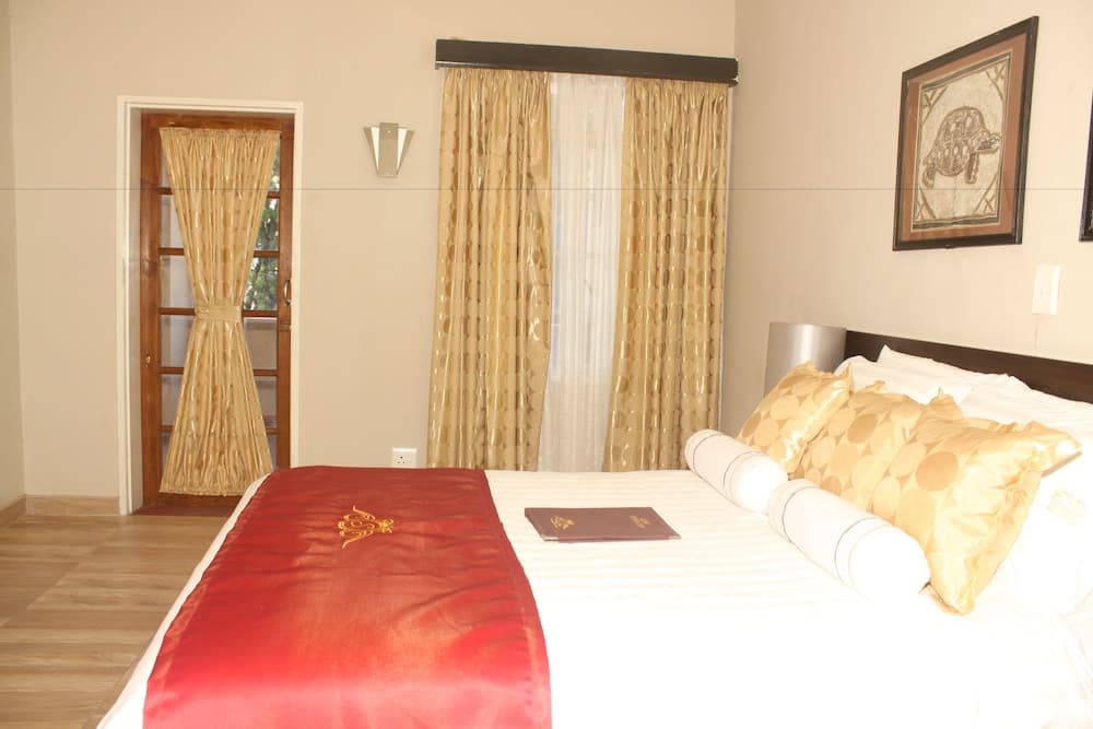 A cozy hotel bedroom features a neatly made bed adorned with golden pillows and a red throw, framed by elegant curtains and a serene view through glass doors, ideal for travelers seeking comfort and relaxation.